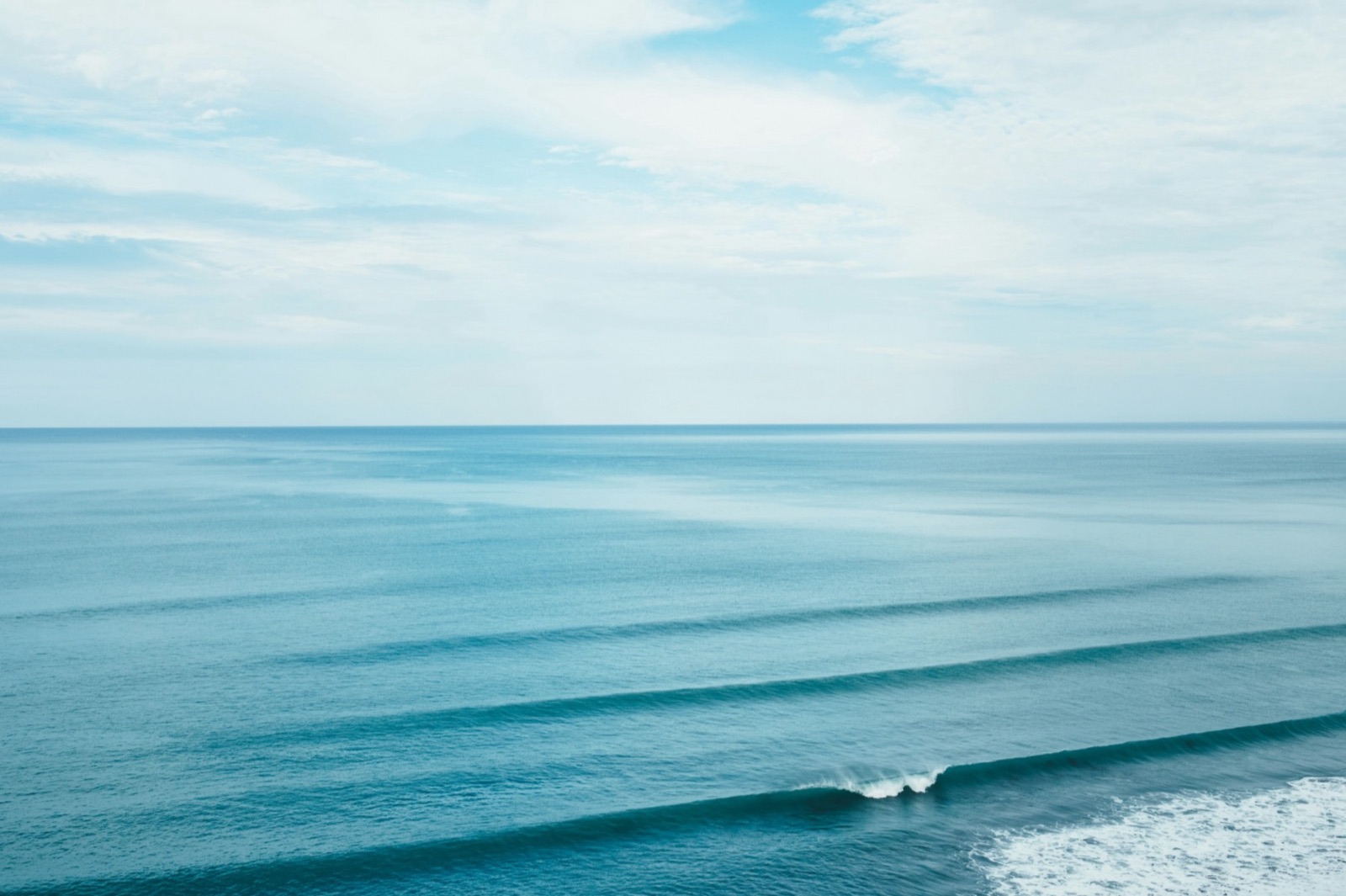 Long, even lines of swell stacking toward the shore under a pale sky.